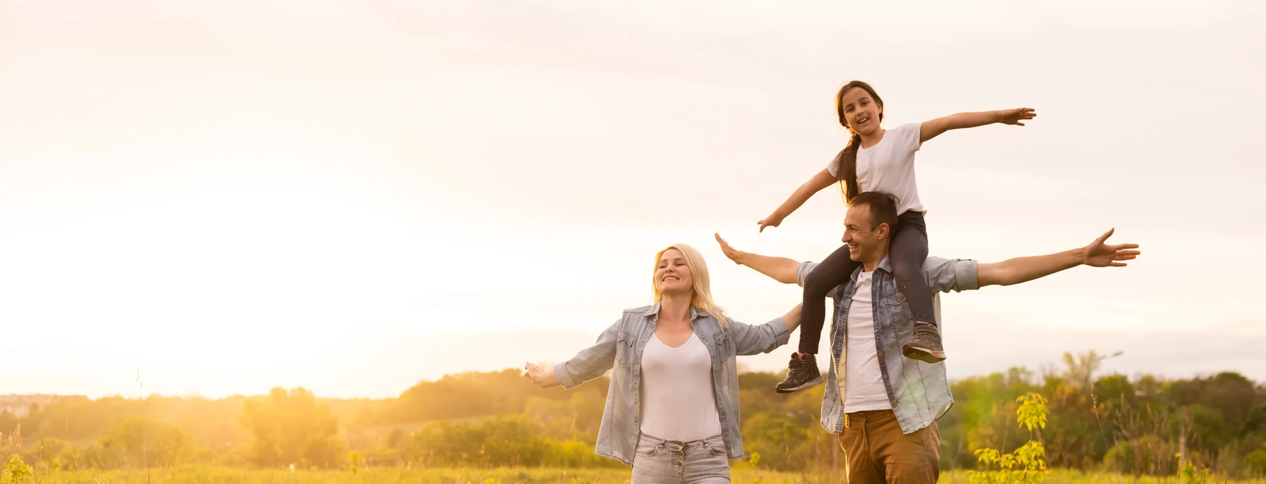 A family of three enjoying a sunny day in a field. The child sits on one adult's shoulders with arms outstretched.