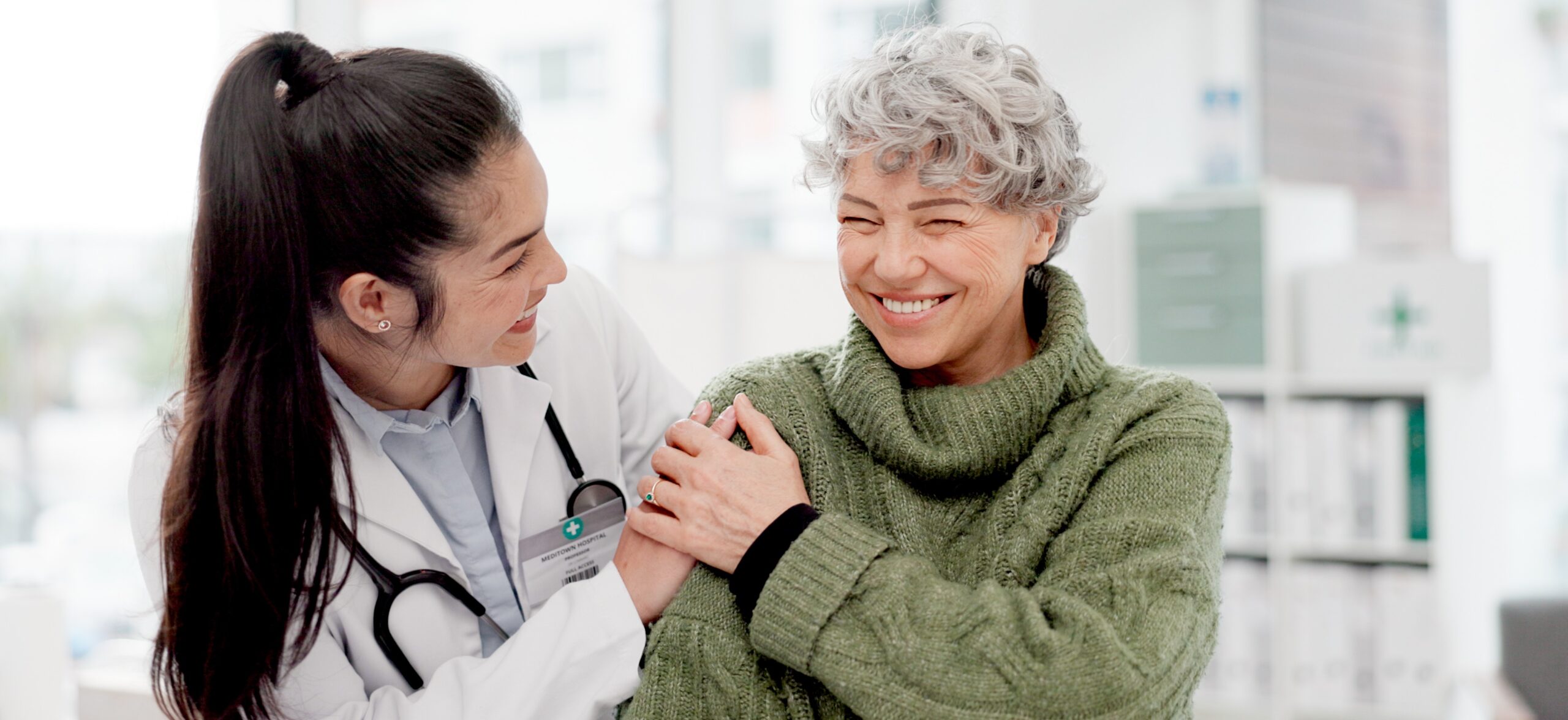A doctor with a stethoscope comforts a smiling elderly woman in a green sweater as they sit together in a bright medical office.
